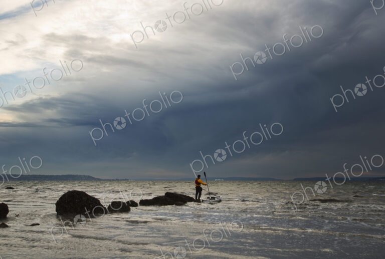 A man stands on a rock in the middle of the Puget Sound with his sea kayak floating…