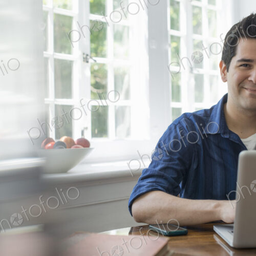 A man using a laptop seated at a table by a window. Working from home. A man using a laptop seated at a table by a window. Working from home.
