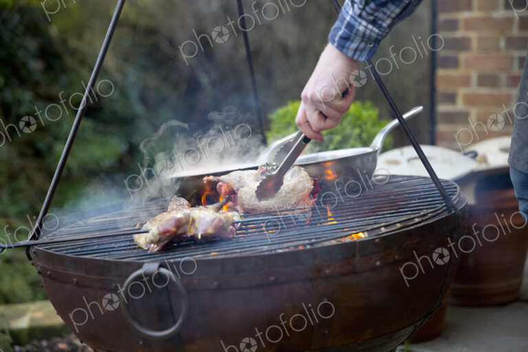 A man using tongs to move roasting game bird pieces on a barbeque.