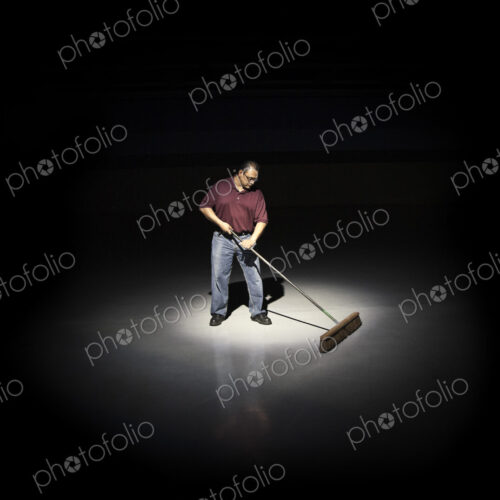 A man with a broom working in an area of a floor lit up by a spotlight.