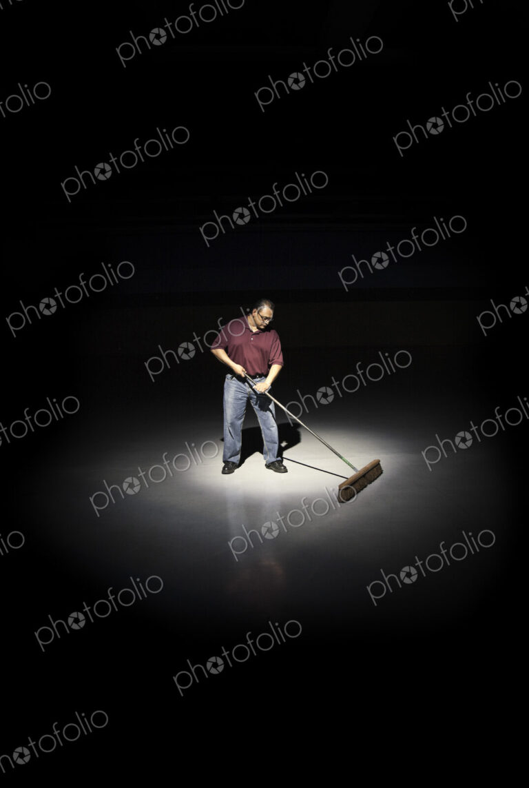 A man with a broom working in an area of a floor lit up by a spotlight.