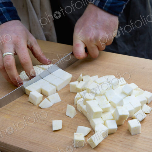 A man with a long knife chopping celeriac into cubes on a chopping board. A man with a long knife chopping celeriac into cubes on a chopping board.