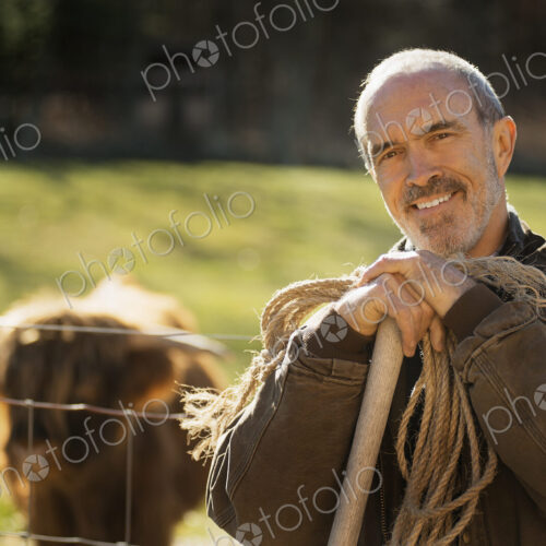 A man with a wooden staff and a coil of rope beside an animal pen. A highland cow with… A man with a wooden staff and a coil of rope beside an animal pen. A highland cow with…
