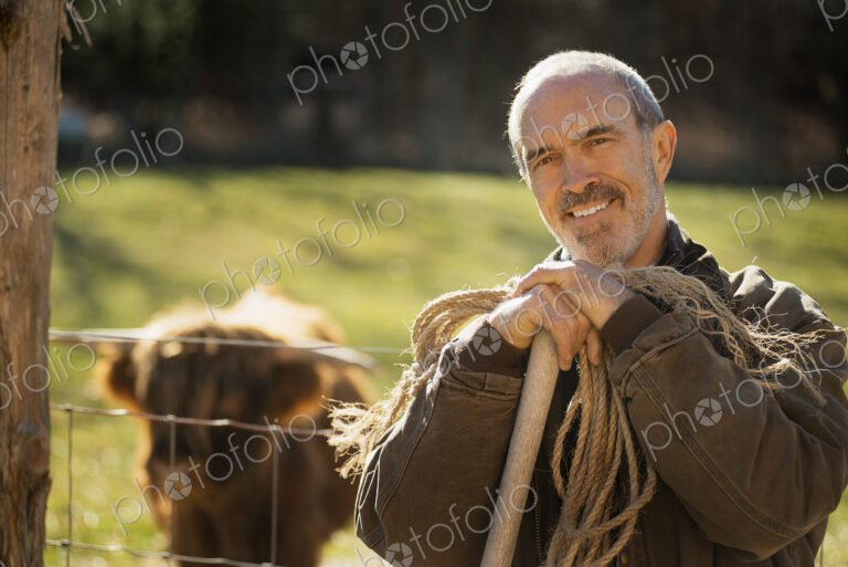 A man with a wooden staff and a coil of rope beside an animal pen. A highland cow with…