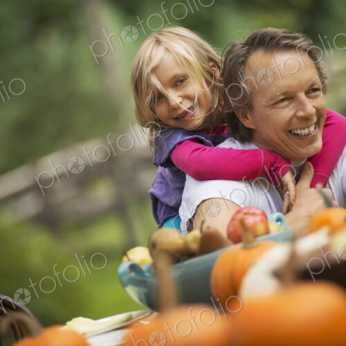 A man with a young girl hugging him from behind, sitting outdoors at a harvest table. A man with a young girl hugging him from behind, sitting outdoors at a harvest table.