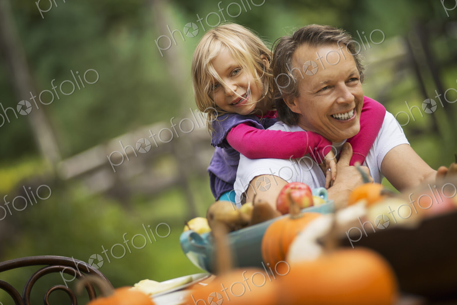 A man with a young girl hugging him from behind, sitting outdoors at a harvest table.