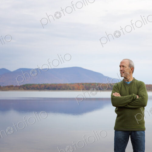 A man with arms folded by the water of a flat calm lake in the mountains.