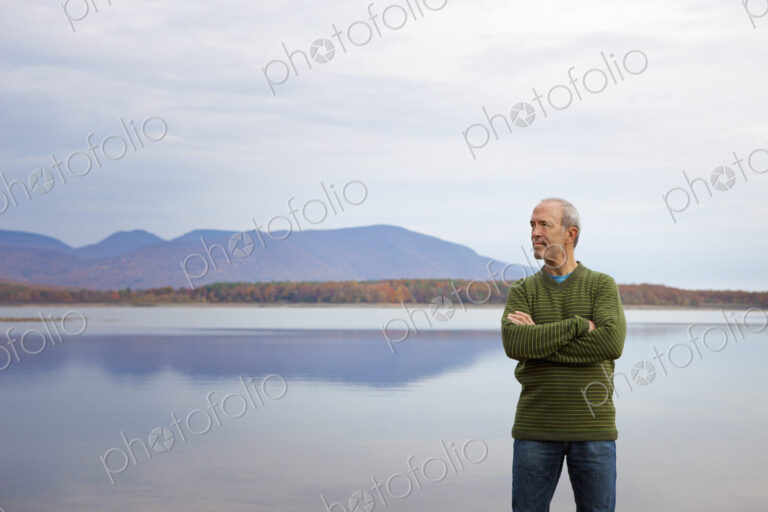 A man with arms folded by the water of a flat calm lake in the mountains.
