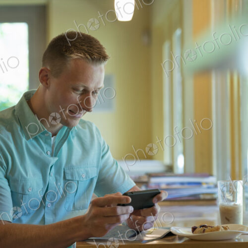 A man with short cropped hair sitting at a cafe table, using a smart phone.