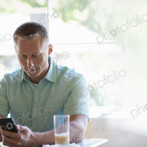 A man with short cropped hair sitting at a cafe table, using a smart phone.