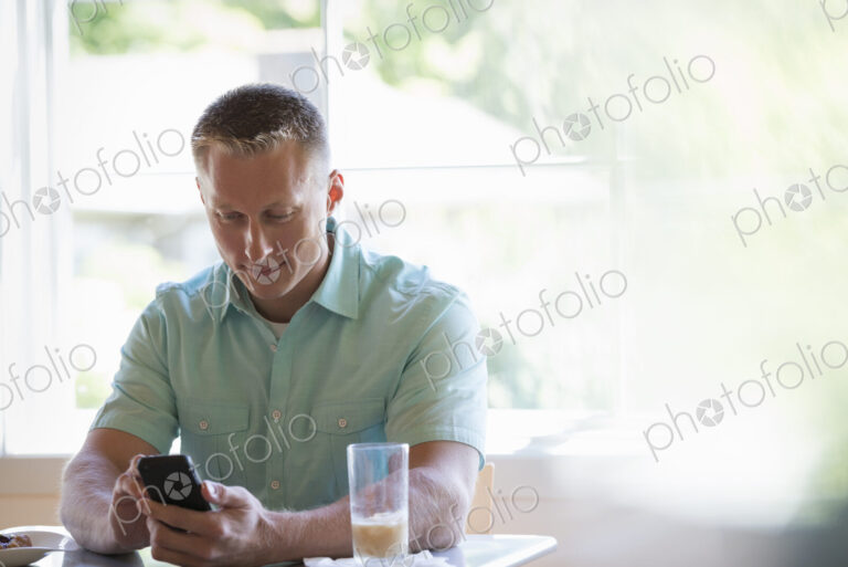 A man with short cropped hair sitting at a cafe table, using a smart phone.