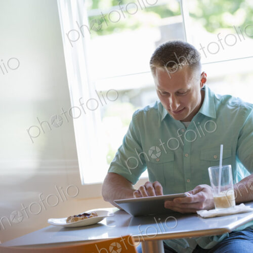A man with short cropped hair sitting at a cafe table. Using a digital tablet.