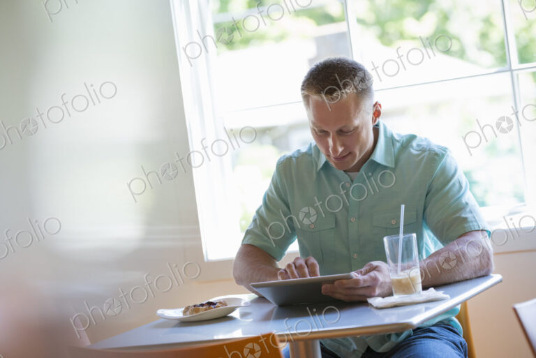 A man with short cropped hair sitting at a cafe table. Using a digital tablet.