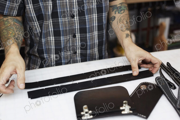 A man with tattoed arms, craftsman working at a bench, smoothing strips of tanned…