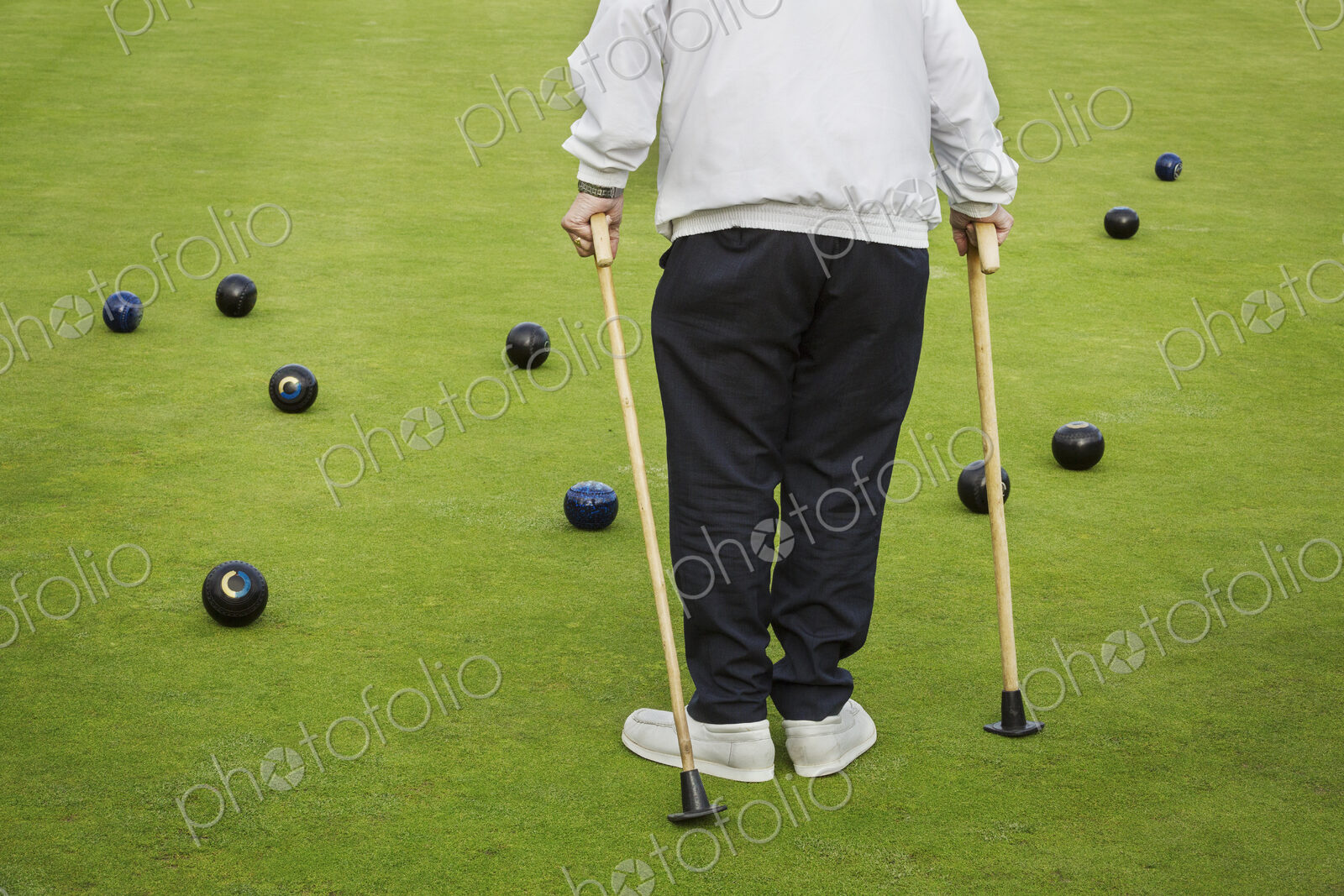 A man with two walking sticks standing on a lawn bowls green, with bowls balls…