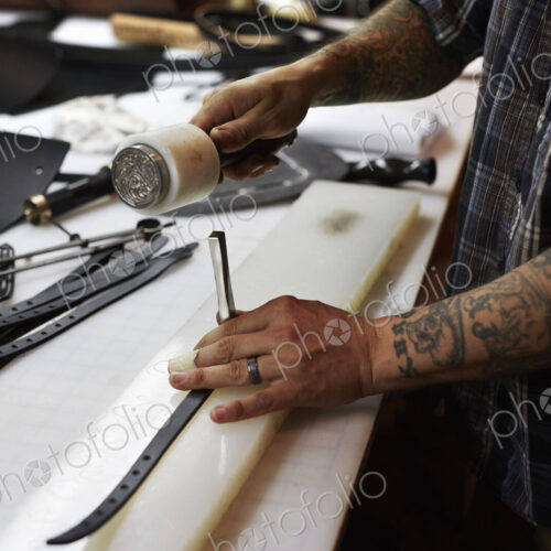A man working at a bench in a leather workshop, using a mallet and chisel to mark leather. A man working at a bench in a leather workshop, using a mallet and chisel to mark leather.