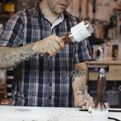 A man working at a bench in a leather workshop, using a mallet and chisel to mark leather. A man working at a bench in a leather workshop, using a mallet and chisel to mark leather.
