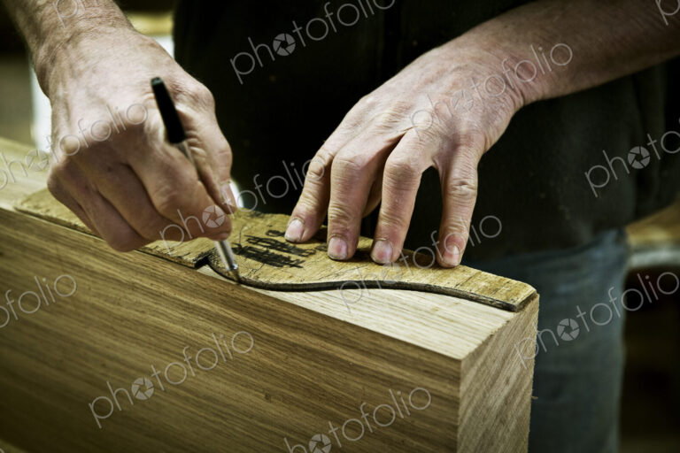 A man working in a furniture maker's workshop drawing around a shape on wood.