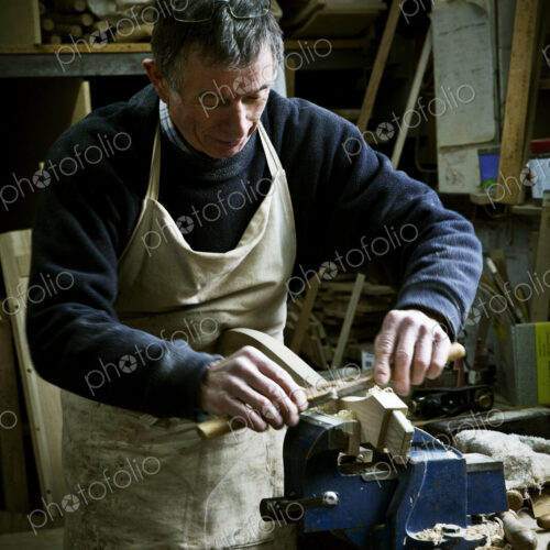 A man working in a furniture maker's workshop, sanding a piece of wood held in a clamp.