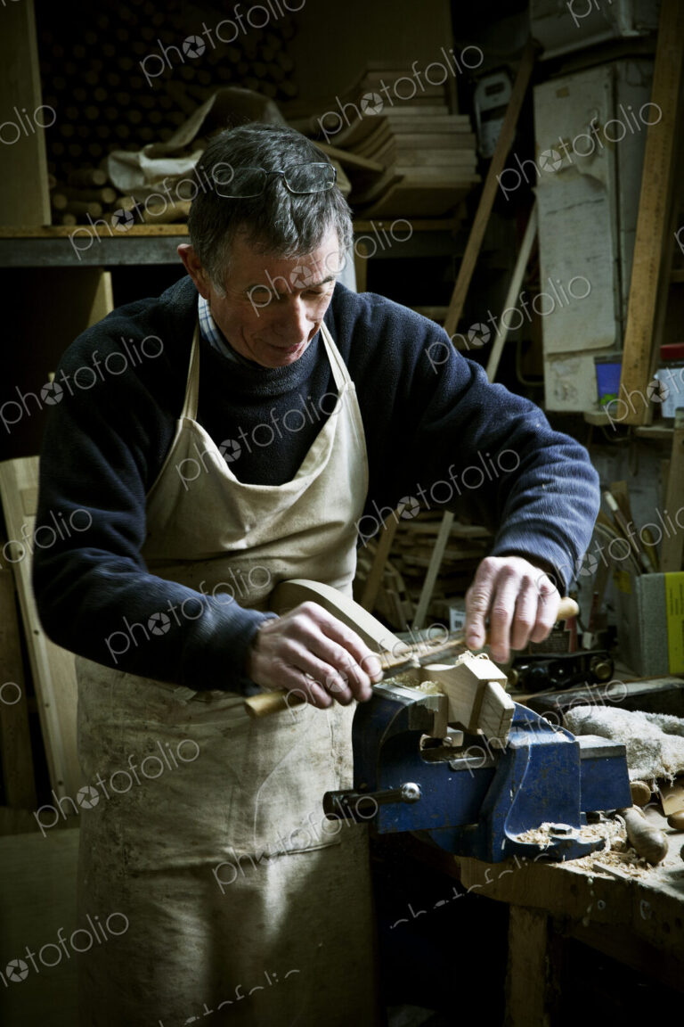 A man working in a furniture maker's workshop, sanding a piece of wood held in a clamp.