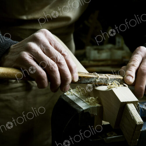 A man working in a furniture maker's workshop, using a rasp on a piece of wood in a clamp.