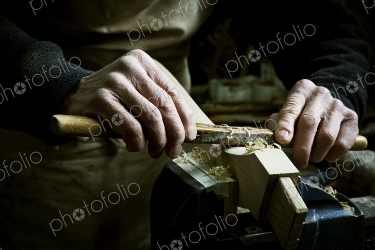 A man working in a furniture maker's workshop, using a rasp on a piece of wood in a clamp.