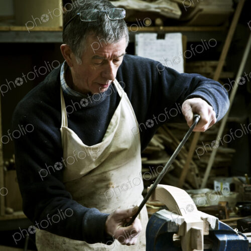 A man working in a furniture maker's workshop using a rasp on a wooden shape in a clamp. A man working in a furniture maker's workshop using a rasp on a wooden shape in a clamp.
