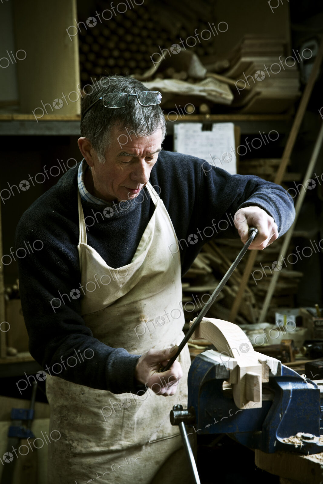 A man working in a furniture maker’s workshop using a rasp on a wooden shape in a clamp.