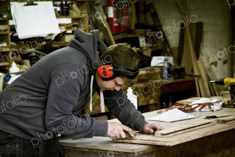 A man working in a furniture maker's workshop wearing ear defenders and using a sharp…