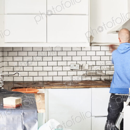 A man working in a new kitchen, a tiler applying tiles to the wall behind the cooker. A man working in a new kitchen, a tiler applying tiles to the wall behind the cooker.