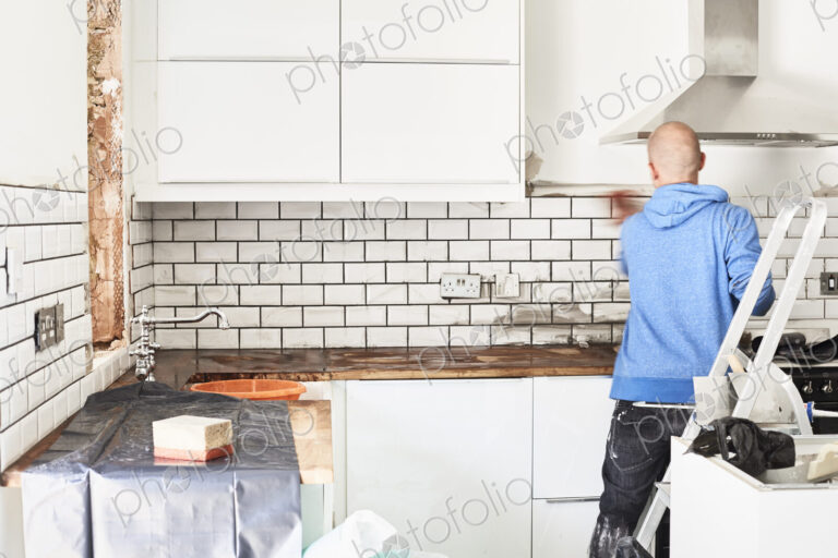 A man working in a new kitchen, a tiler applying tiles to the wall behind the cooker.