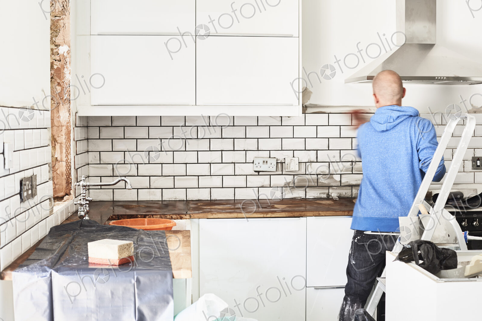 A man working in a new kitchen, a tiler applying tiles to the wall behind the cooker.