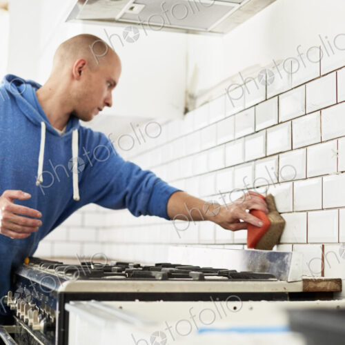 A man working in a new kitchen, a tiler applying tiles to the wall behind the cooker. A man working in a new kitchen, a tiler applying tiles to the wall behind the cooker.