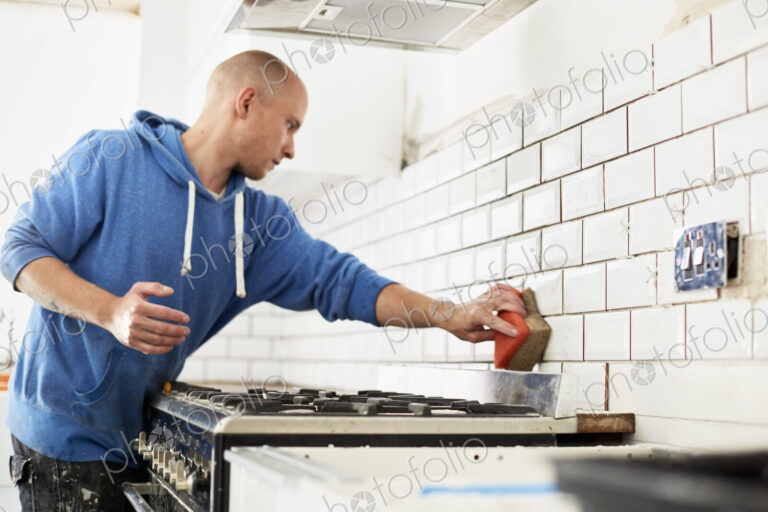 A man working in a new kitchen, a tiler applying tiles to the wall behind the cooker.