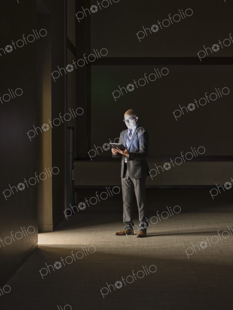 A mature Asian businessman standing in shadows in an office building, looking at a…
