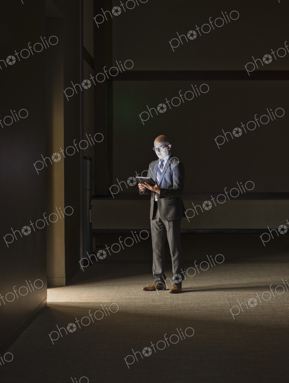 A mature Asian businessman standing in shadows in an office building, looking at a…
