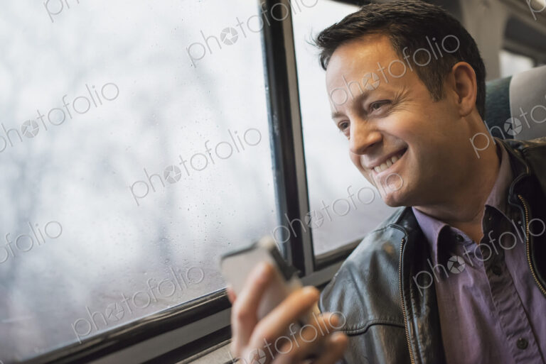 A mature man sitting at a window seat on a train, holding his mobile phone. Smiling…