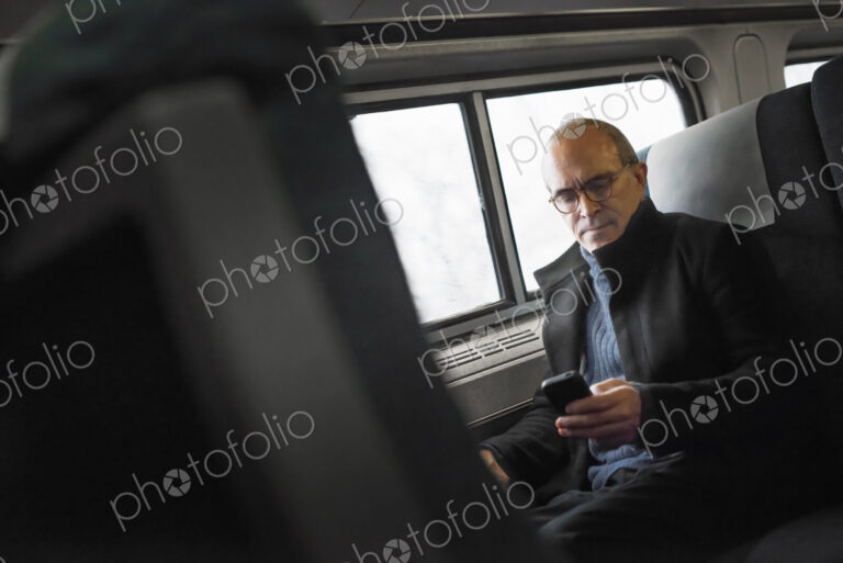 A mature man sitting by a window in a train carriage, using his mobile phone, keeping…