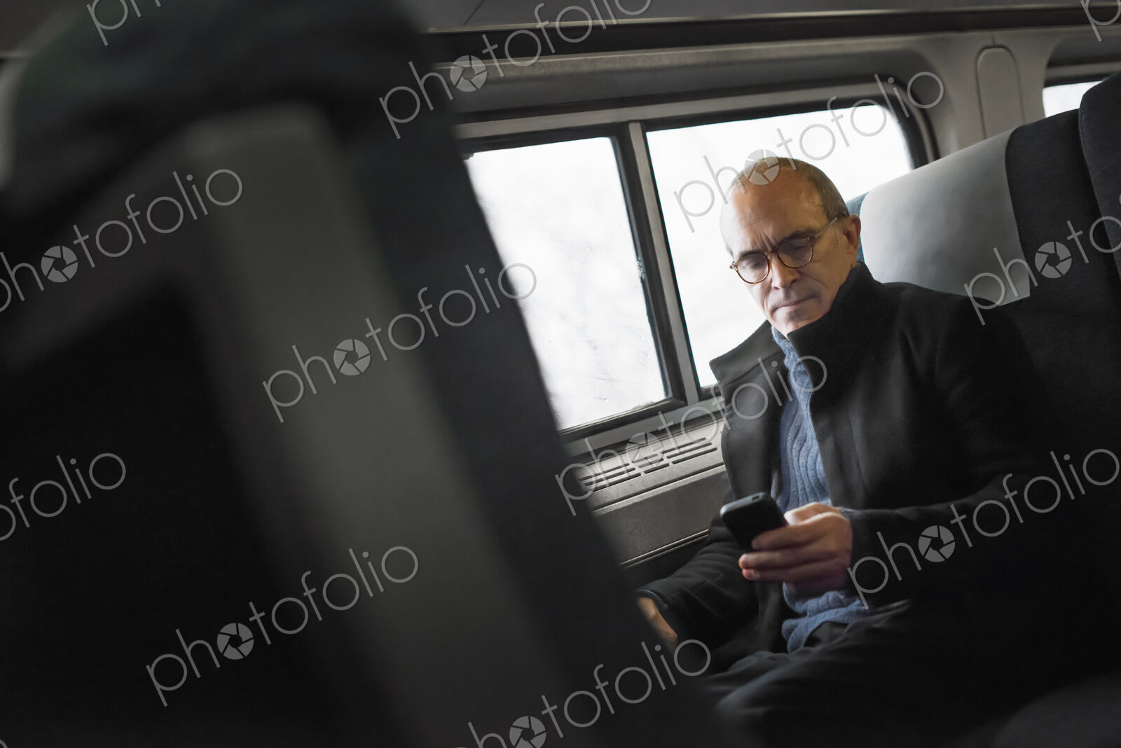 A mature man sitting by a window in a train carriage, using his mobile phone, keeping…