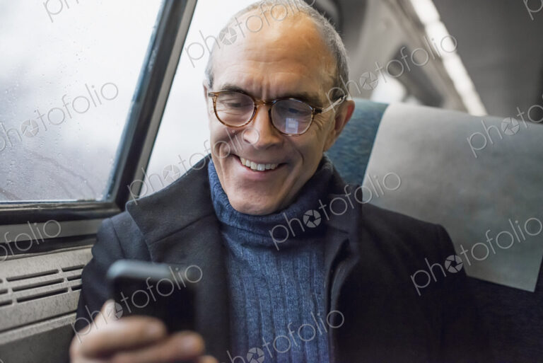 A mature man sitting by a window in a train carriage, using his mobile phone, keeping…