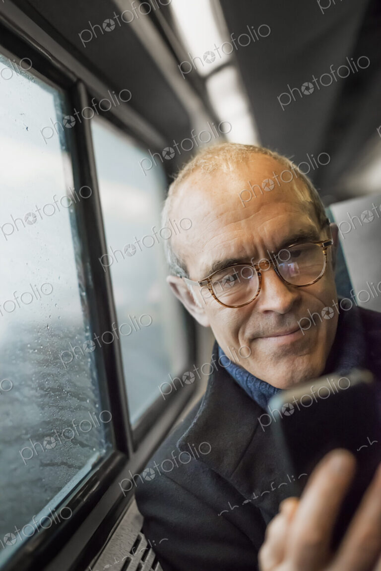 A mature man sitting by a window in a train carriage, using his mobile phone, keeping…