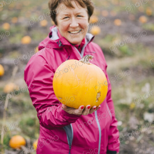 A mature woman in a red jacket, holding out a large orange pumpkin.