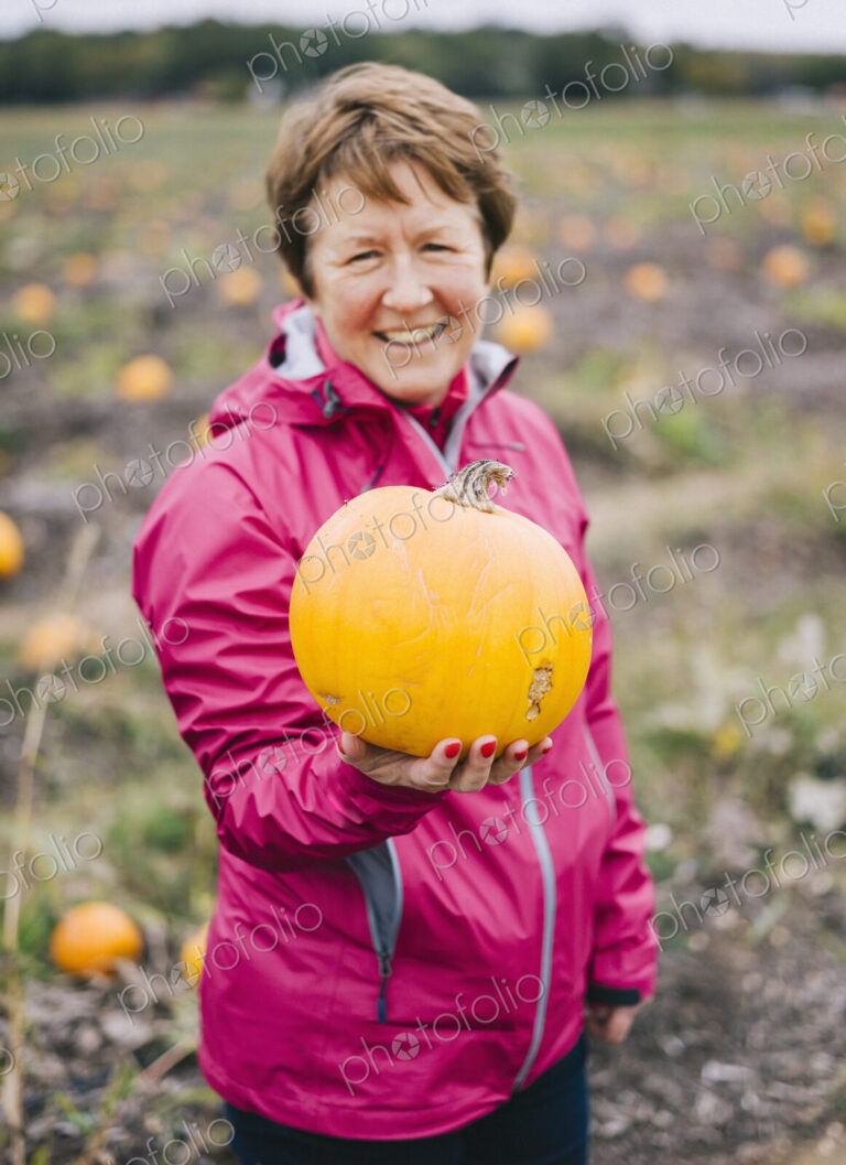 A mature woman in a red jacket, holding out a large orange pumpkin.