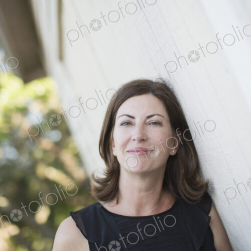 A mature woman in a sleeveless shirt with shoulder length brunette hair by a white wall.