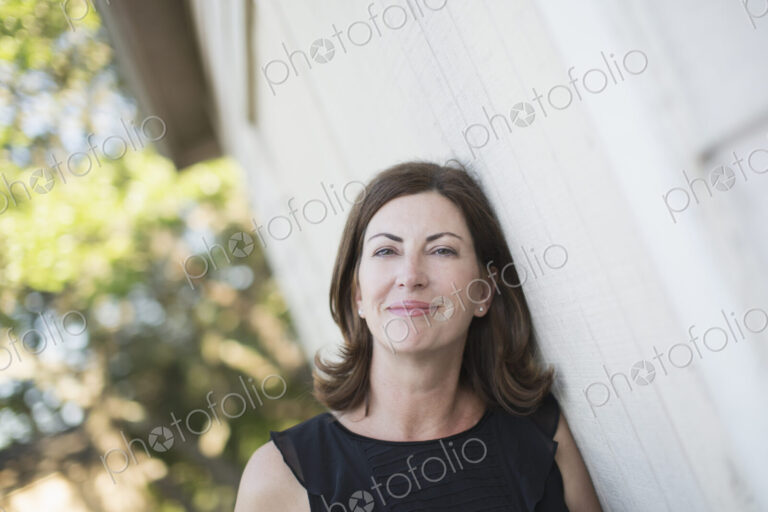 A mature woman in a sleeveless shirt with shoulder length brunette hair by a white wall.