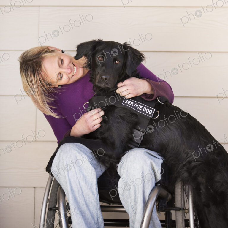 A mature woman wheelchair user with her arms around her service dog, a black labrador…