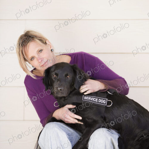 A mature woman wheelchair user with her arms around her service dog, a black labrador…
