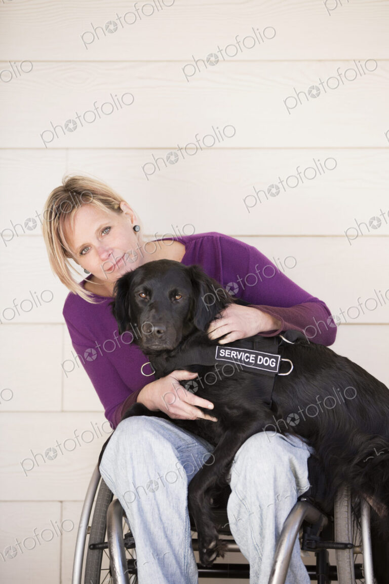 A mature woman wheelchair user with her arms around her service dog, a black labrador…
