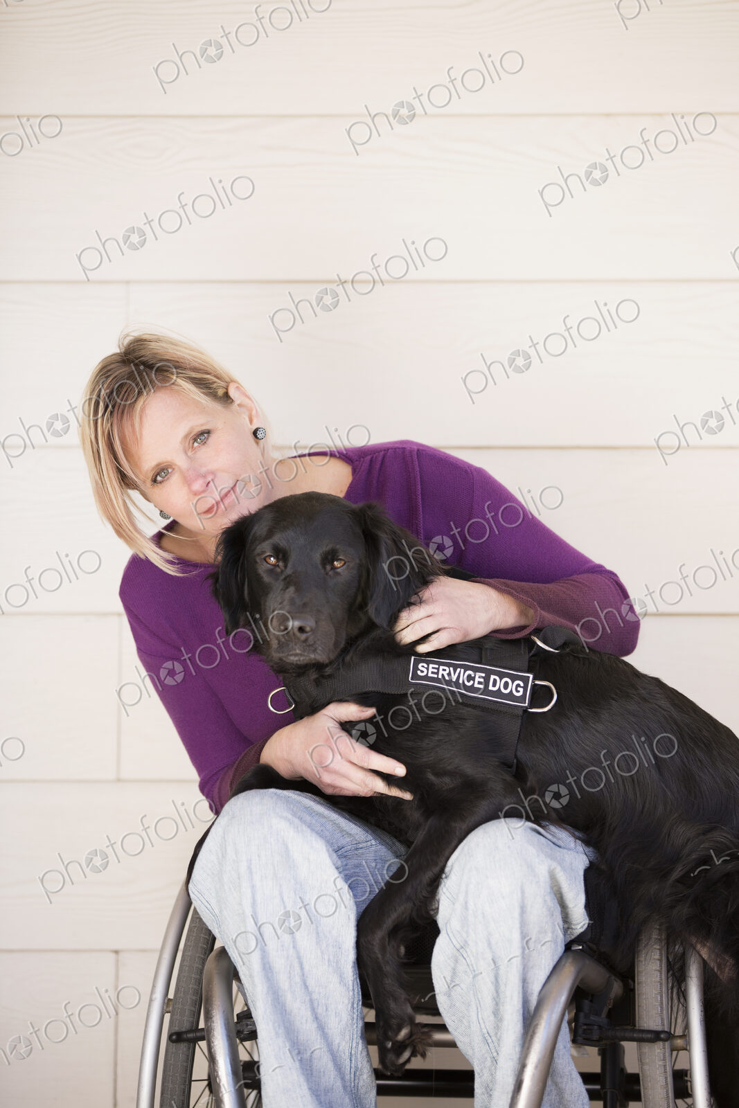 A mature woman wheelchair user with her arms around her service dog, a black labrador…