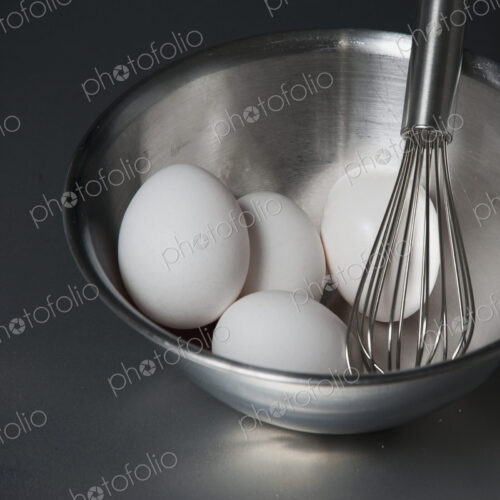 A metal bowl with a balloon whisk and white eggs.